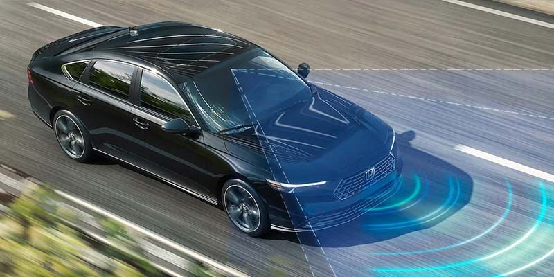 Overhead view of a black sedan driving on a highway, with a visualized driver-assistance or collision detection system highlighted in blue ahead of the vehicle.