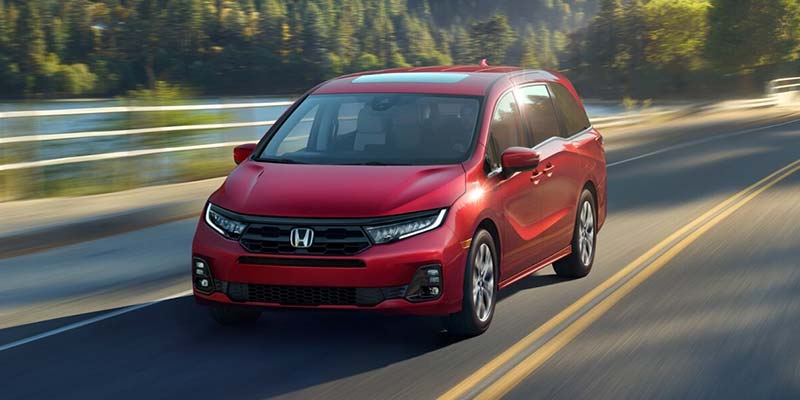 Front three-quarter view of a red Honda SUV driving on a sunlit road, with motion blur in the background emphasizing speed and a scenic wooded landscape.