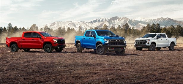 Chevy pickup trucks lined up on an off-road dirt field with dust in the air, set against a mountain backdrop with snow-capped peaks, showcasing red, blue, and white truck variants.