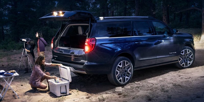 Dark blue Ford Expedition with the rear hatch open at a campsite, showing people preparing supplies and using a telescope at dusk