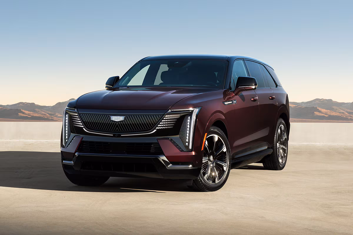 Front three-quarter view of a maroon Cadillac Escalade IQ parked in an open landscape with mountains in the background, highlighting its exterior design and wheels.