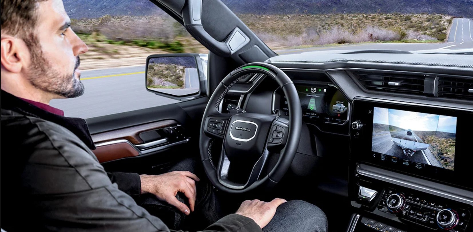 Driver inside a GMC truck towing a trailer while viewing the rear camera feed on the infotainment screen, with highway and landscape visible outside.