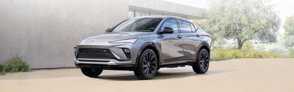 2025 Buick Envista in sleek silver parked in a modern driveway, showcasing its aerodynamic design, black alloy wheels, and stylish front grille.