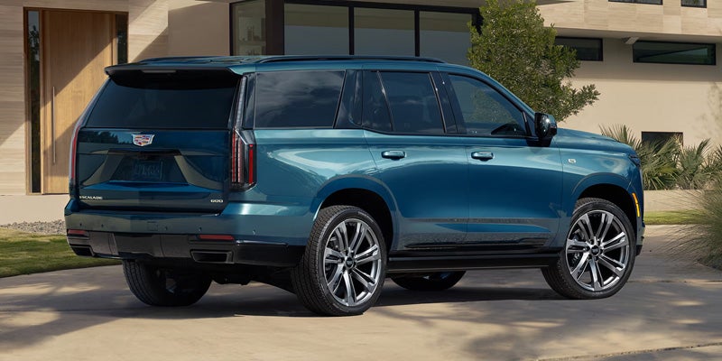 A rear-three-quarter view of a dark teal Cadillac Escalade parked on a concrete driveway in front of a modern, multi-story home.