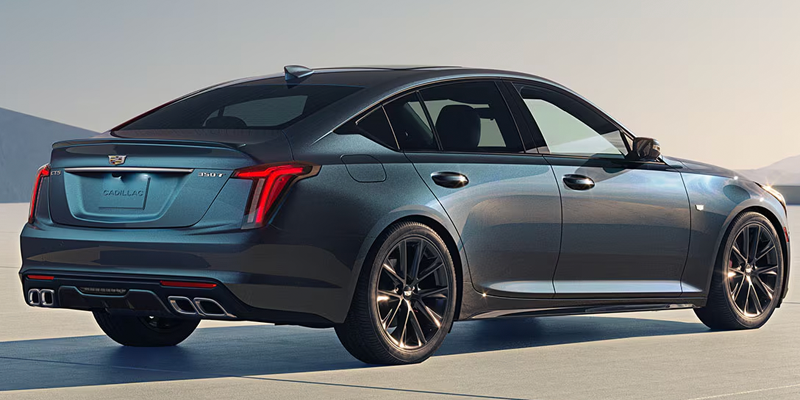 Rear-three-quarter view of a dark metallic blue-gray Cadillac CT5 350T sedan parked outdoors on an open paved surface against a clear sky.