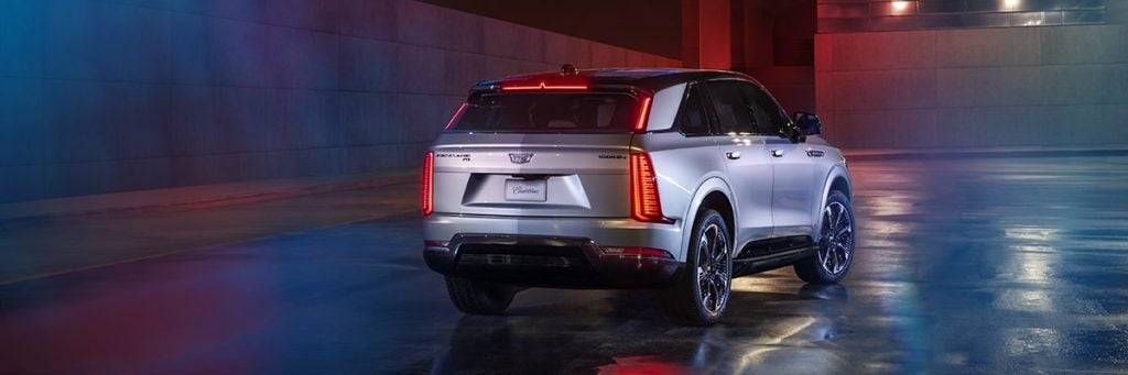 Rear-three-quarter view of a silver Cadillac Escalade IQ parked in a modern, neon-lit concrete area at night.
