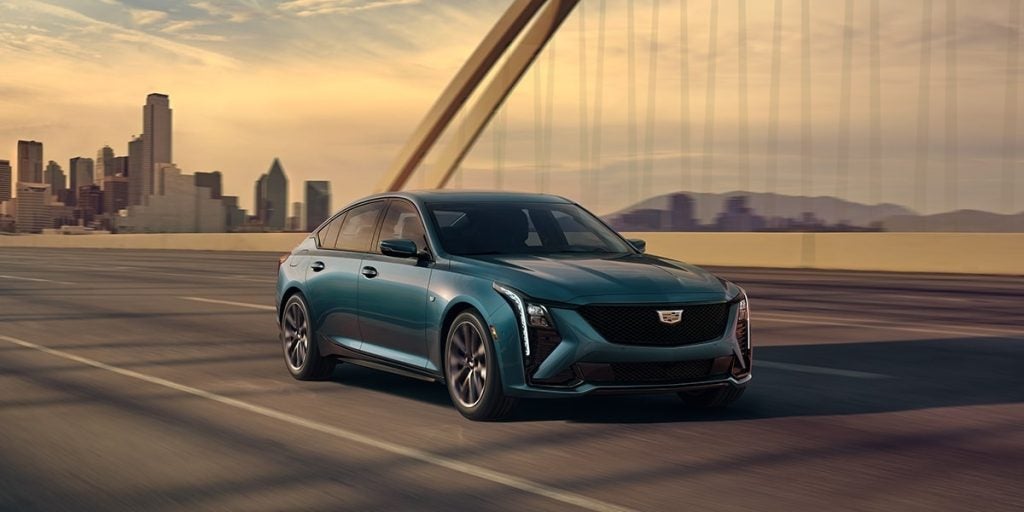 Front-three-quarter view of a teal Cadillac CT5 sedan driving across a large suspension bridge with a city skyline in the background at dusk.