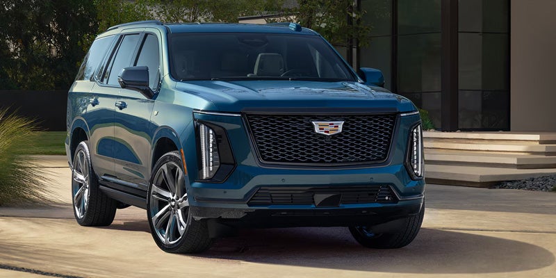 Front-three-quarter view of a dark teal Cadillac Escalade SUV parked in a driveway outside a modern home with large glass windows.