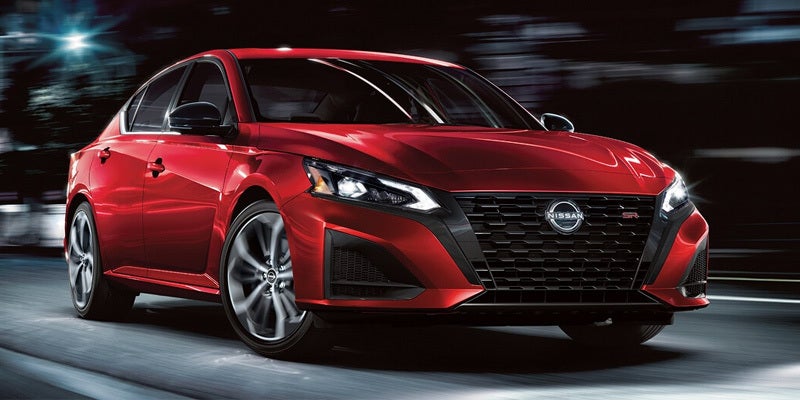 Front three-quarter view of a bright red Nissan Altima SR sedan driving down a city street at night with motion-blurred city lights in the background.