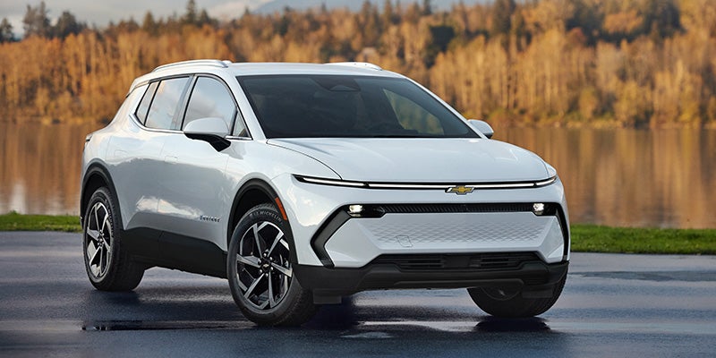 White Chevrolet Equinox EV parked near a lake with autumn trees in the background.