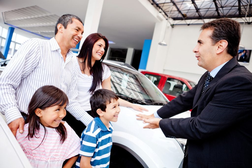 Family at a car dealership interacting with a salesperson while viewing a vehicle, with parents and children smiling during the car-buying experience.