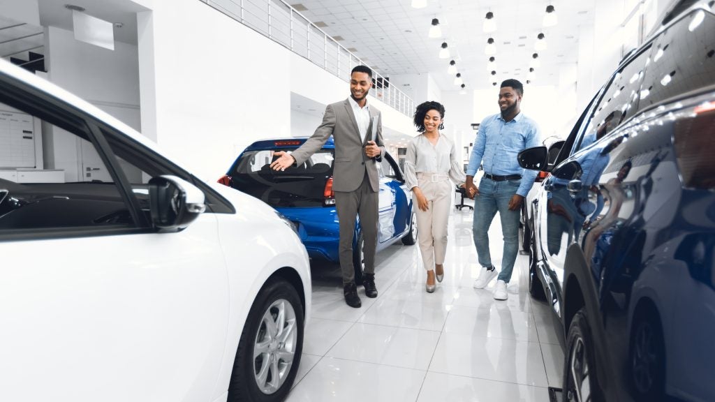 Three people walking through a car dealership showroom while a salesperson presents vehicles, with cars displayed on both sides in a bright modern interior.