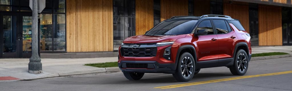 A vibrant red 2025 Chevrolet Equinox with a sleek black roof and silver trimmings approaches a stop sign in downtown Seminole, OK, during daylight. To its right, a modern corner building with a warm brown wood exterior, black accents, and large glass windows adds to the urban backdrop.