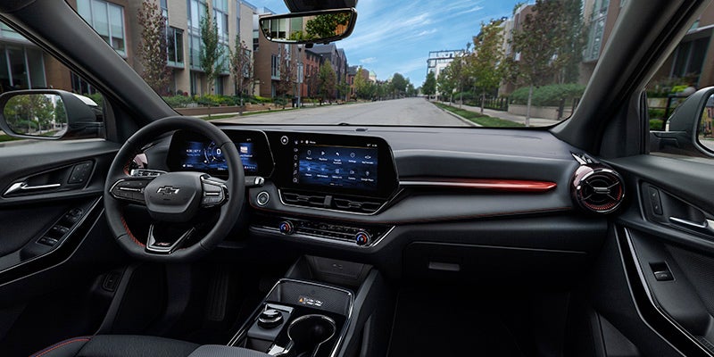 A passenger-side view of the 2025 Chevrolet Equinox, parallel parked along a sidewalk on a neighborhood road lined with apartments near Seminole, OK. The interior features a sleek black and gray color scheme with leather seats. The infotainment screen and digital dash display are illuminated, while red ambient lighting adds a sophisticated touch to the passenger-side dashboard.