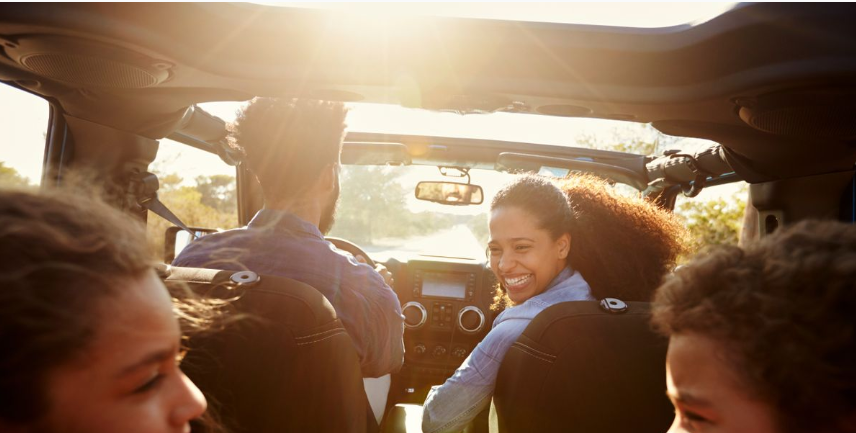 Passengers enjoying a sunny drive in a convertible SUV with the top down, smiling and looking back at the camera with sunlight streaming into the cabin.