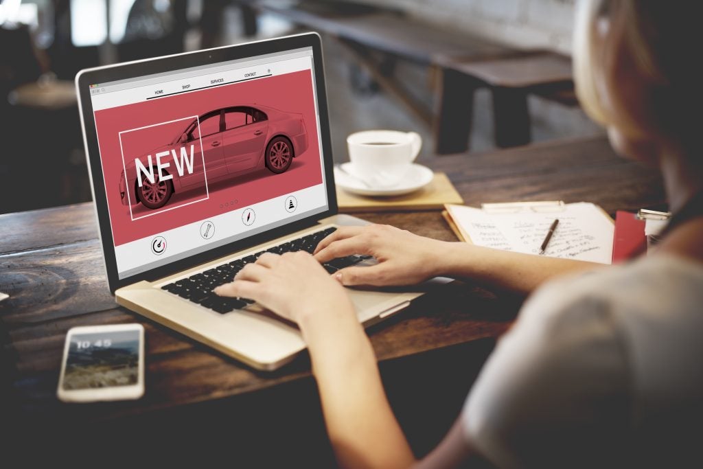 A person browsing a car website on a laptop in a café, with a "NEW" car advertisement on the screen.

