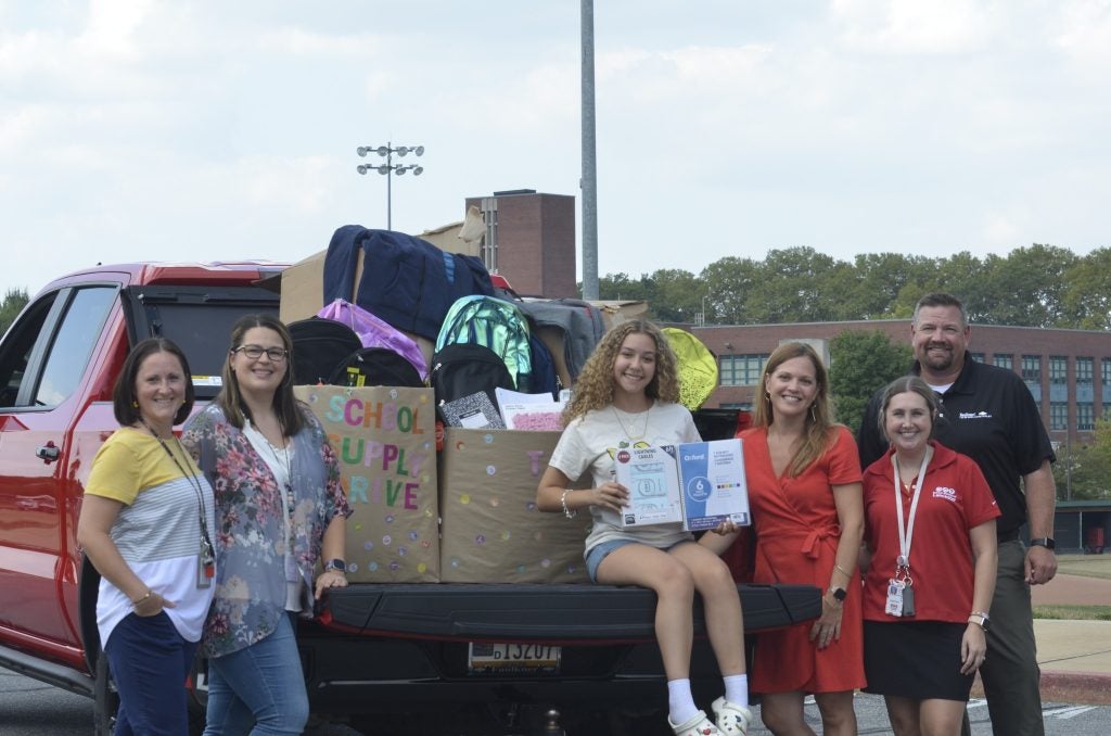 a group of people standing around the back of a truck, which has been loaded with bags full of school supplies.