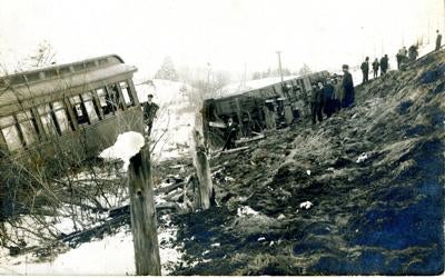 Historical photograph showing onlookers gathered around the wreckage of derailed train cars from the 1907 St. John &amp; Lake Champlain Railroad accident, with damaged passenger cars visible at the bottom of the embankment