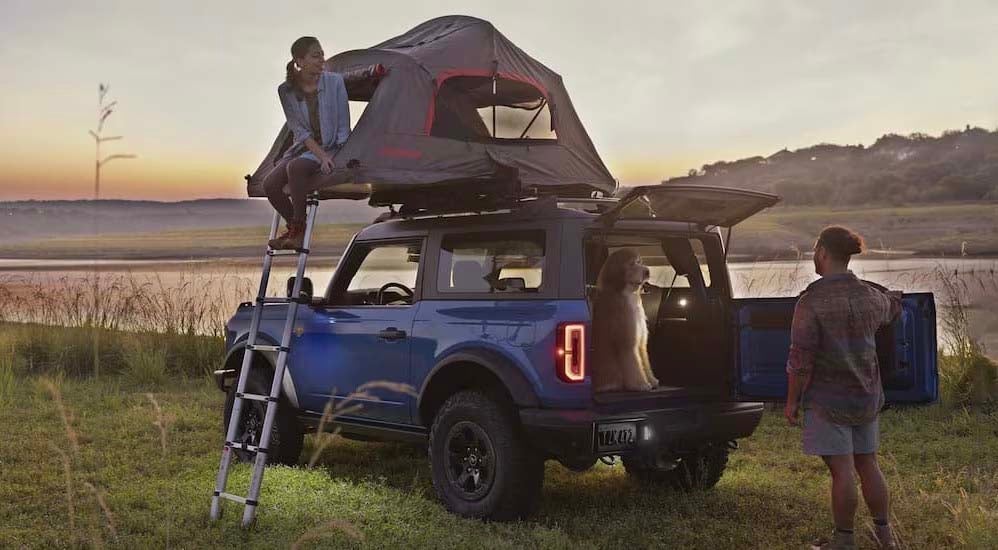A blue 2024 Ford Bronco is shown parked off-road near a pond and campers.