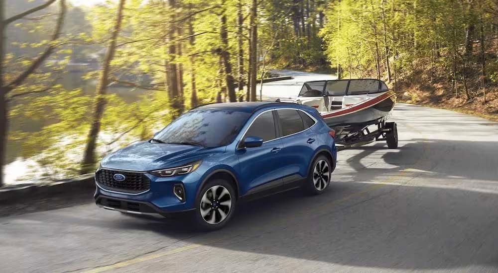 A blue 2023 Ford Escape is shown towing a boat on a winding road next to a lake.