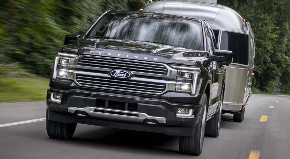 A black 2024 Ford F-150 Hybrid Platinum is towing a grey trailer on a tree-lined road.