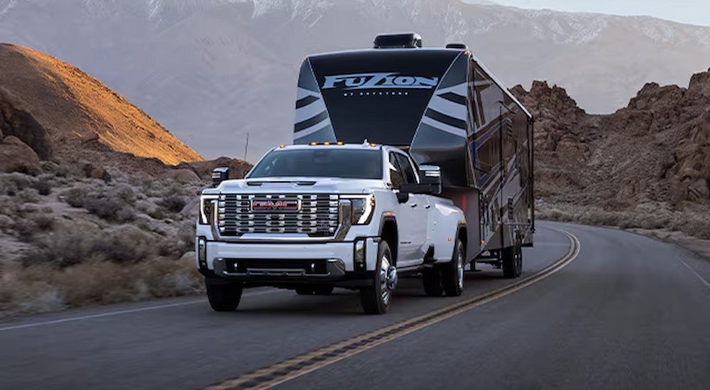 A white 2025 GMC Sierra 3500 HD Denali is towing a camper on an open road after viewing trucks for sale near Minneapolis.