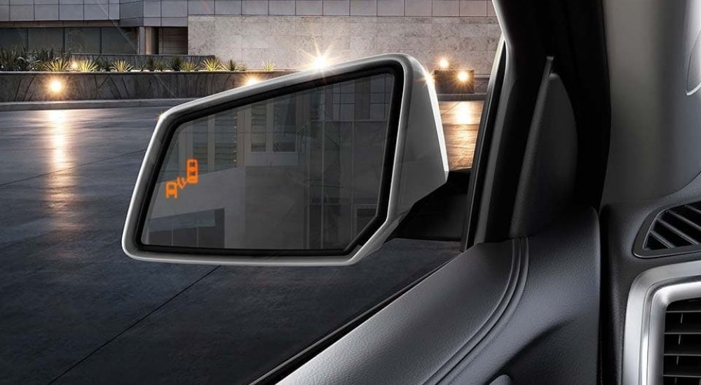 An illuminated blind spot monitoring icon is shown on the mirror of a silver 2015 GMC Acadia.