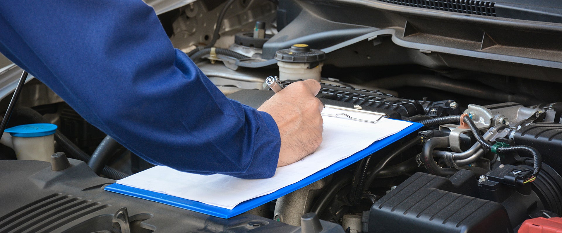 A mechanic filling out a checklist on a clipboard over an open car engine bay.