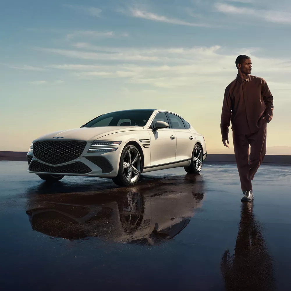 White Genesis G80 luxury sedan parked on a reflective wet surface, with a man walking beside it.