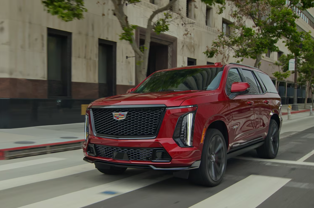A sleek red Cadillac Escalade V with a prominent grille and sharp headlights drives down a city street. The scene conveys a sense of power and elegance.