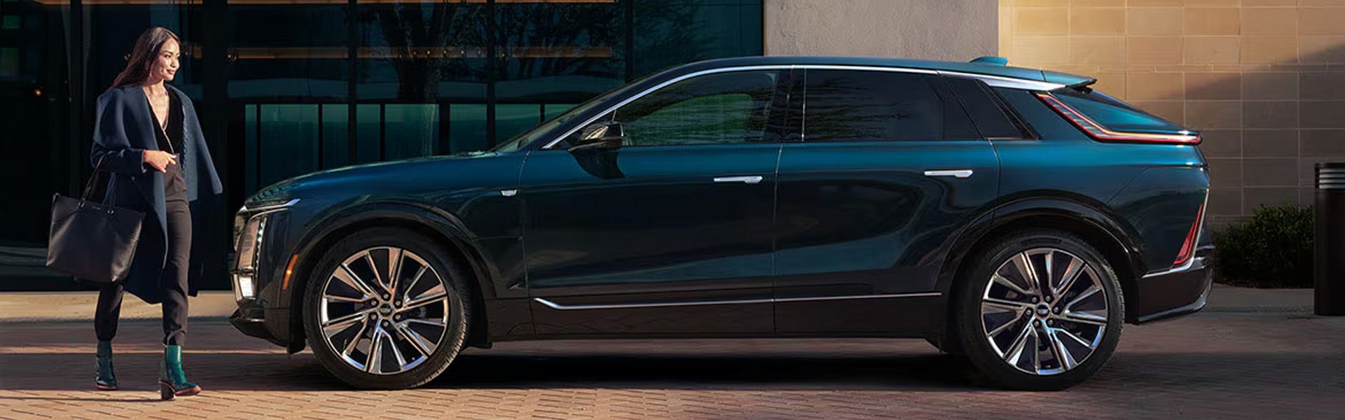 A sleek 2026 Cadillac LYRIQ electric SUV in dark metallic blue parked in front of a modern building, with a well-dressed woman walking beside it carrying a large handbag.