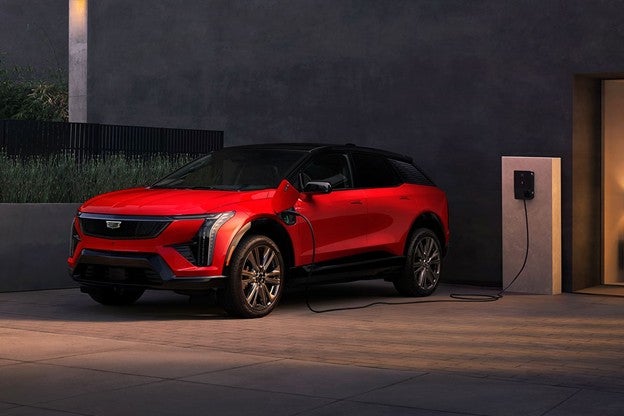 Red 2026 Cadillac XT5 parked in a modern outdoor setting at night, shown plugged into a wall-mounted charging unit, with sleek LED lighting and a black roof contrasting the red body.