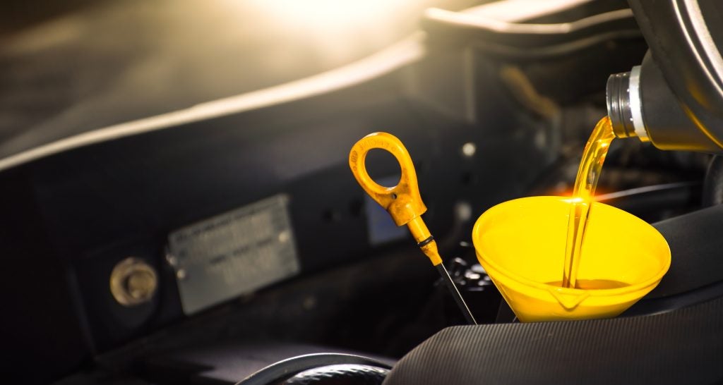 Engine oil being poured into a funnel in a car engine bay, with a dipstick visible nearby.