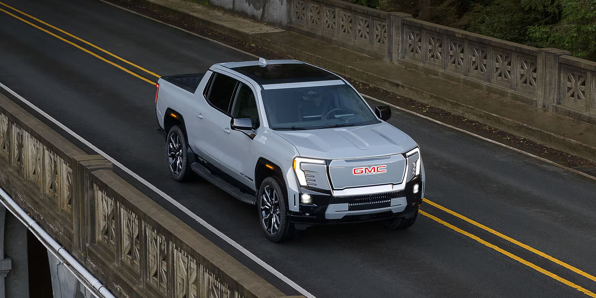 A silver GMC Sierra EV pickup truck driving on a two-lane road beside a stone barrier.