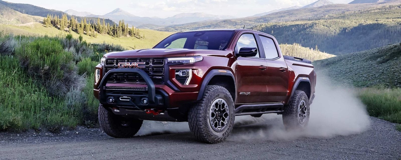 Red 2026 GMC Canyon driving on a gravel road through green hills; new GMC Canyon trucks for sale at Ross Downing GMC in Hammond, LA