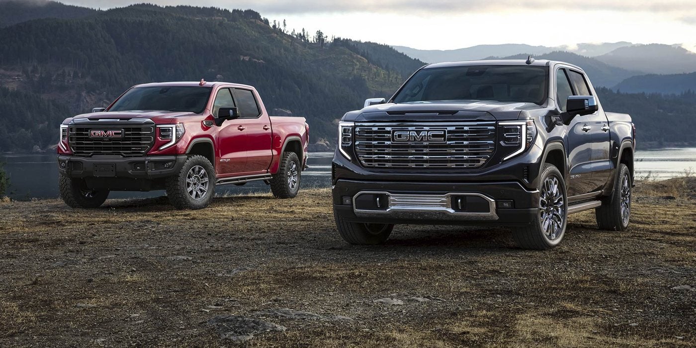 Red 2026 GMC Sierra 1500 AT4 and black 2026 GMC Sierra 1500 Denali parked by a lake, both sold new at Ross Downing GMC in Hammond, LA