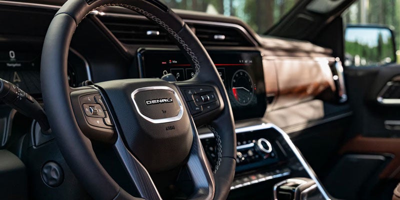 Close-up interior view focusing on a leather-wrapped steering wheel with a chrome "DENALI" emblem, showcasing a digital dashboard screen and premium wood trim in the background.