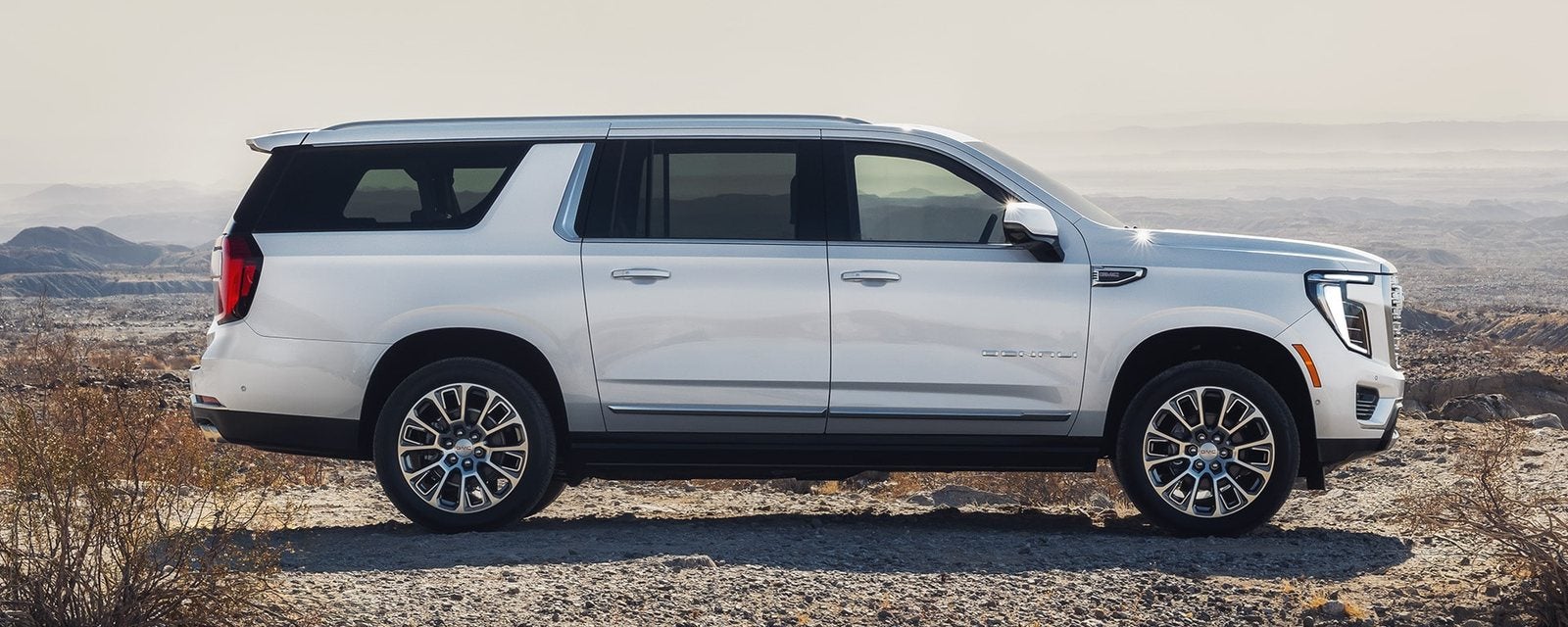 White 2027 GMC Yukon XL in profile on a desert overlook, showing its long body; new GMC Yukon XL SUVs for sale at Ross Downing GMC in Gonzales, LA