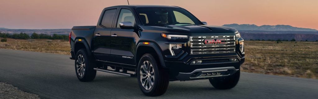 Black GMC pickup truck driving on a road at sunset with mountains in the background.