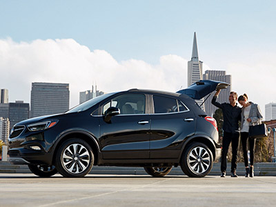 A sleek black SUV is parked with an open trunk. A couple stands nearby, embracing, with a city skyline and skyscrapers in the background.