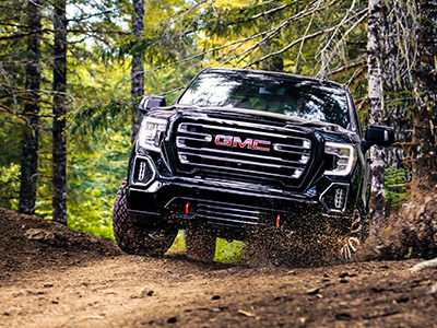 A black GMC truck navigates a rugged, dirt forest trail. Trees surround the path, creating a dynamic, adventurous, and rugged outdoor scene.