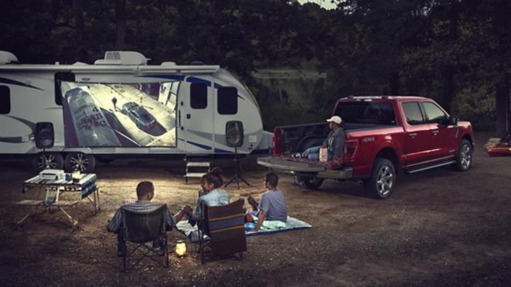a family of four sits and watches a movie thats being projected onto the side of an rv camper