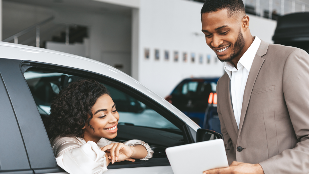 a young woman leans out of a car window smiling as a man points out something on a clipboard to her