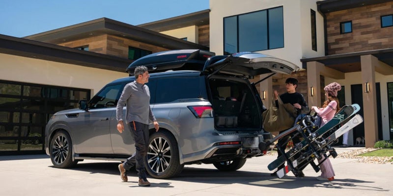 2026 INFINITI QX80 Sport SUV parked outside a modern home in Grapevine, TX, with a family loading ski gear into the rear cargo area, highlighting spaciousness and luxury.
