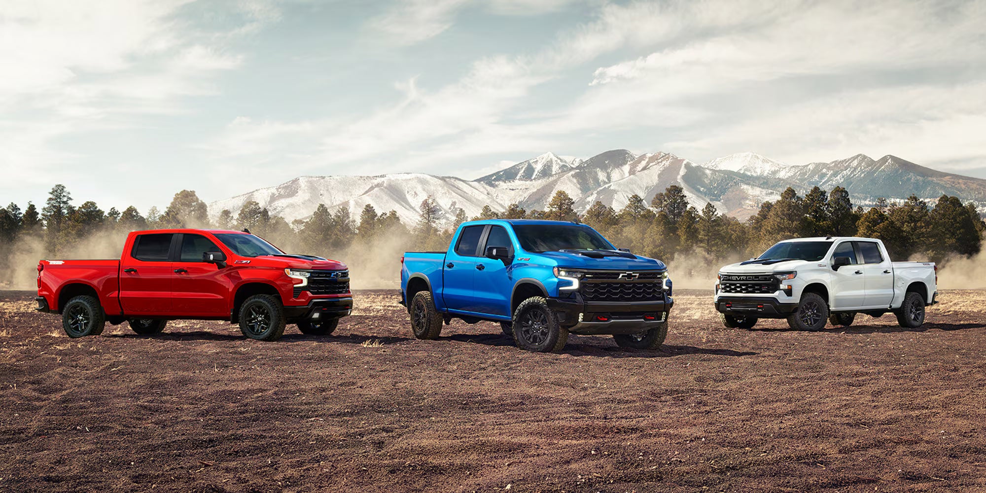 Chevrolet Silverado lineup showcasing a red, blue, and white truck models in a rugged outdoor setting with mountains in the background
