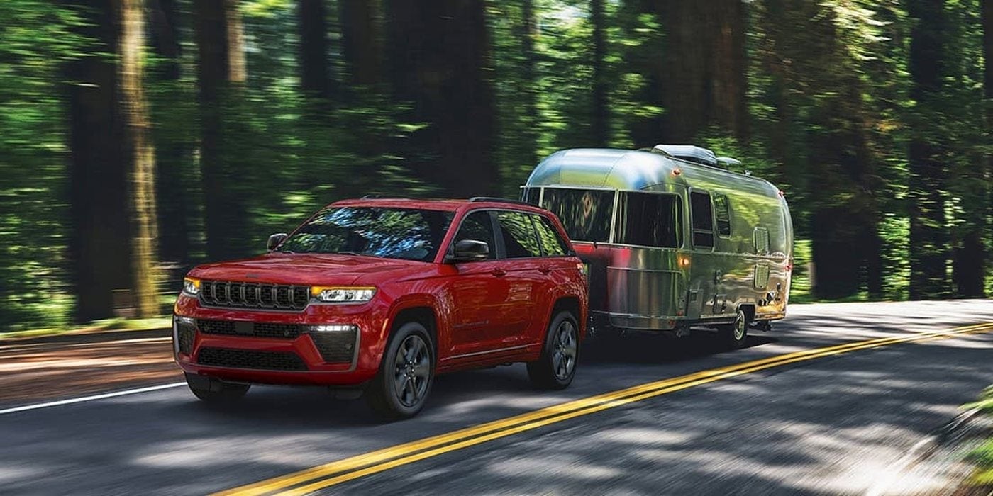 Red 2027 Jeep Grand Cherokee Limited Reserve towing a travel trailer on a wooded highway, for sale at Ross Downing CDJR in Hammond, LA