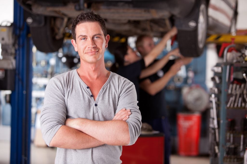 Man inspecting car under lifted vehicle in an auto repair shop, with mechanics working in the background.