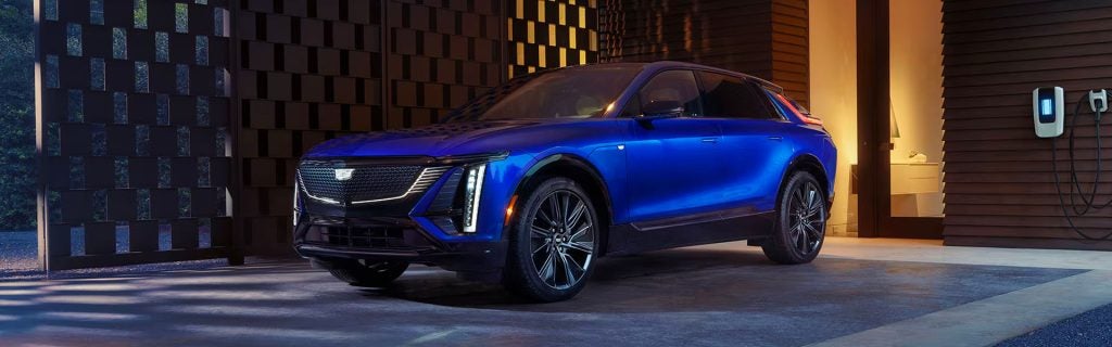 Blue Cadillac LYRIQ electric SUV parked outside a modern home beside a wall-mounted EV charger at night.