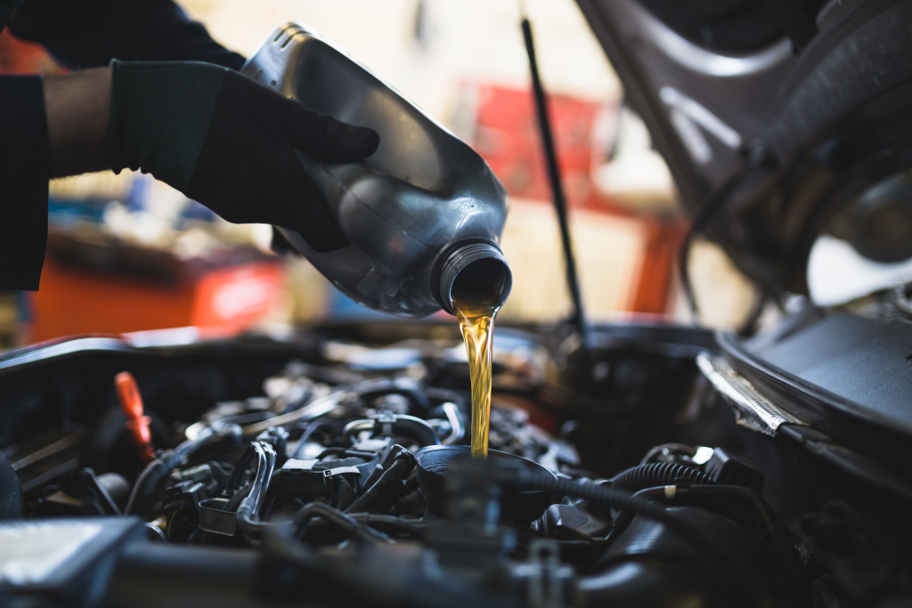Gloved hands pour engine oil from a gray container into a car's open engine bay, conveying precision and maintenance in an automotive setting.