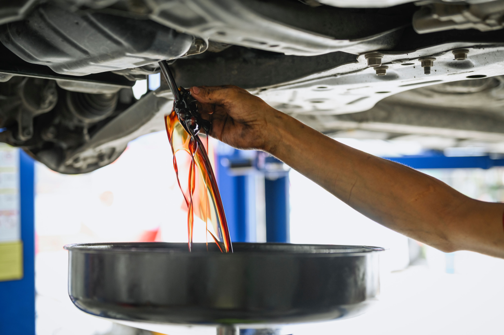 Mechanic's hand draining amber car oil into a black pan under a vehicle. The setting appears to be a well-lit auto repair shop.
